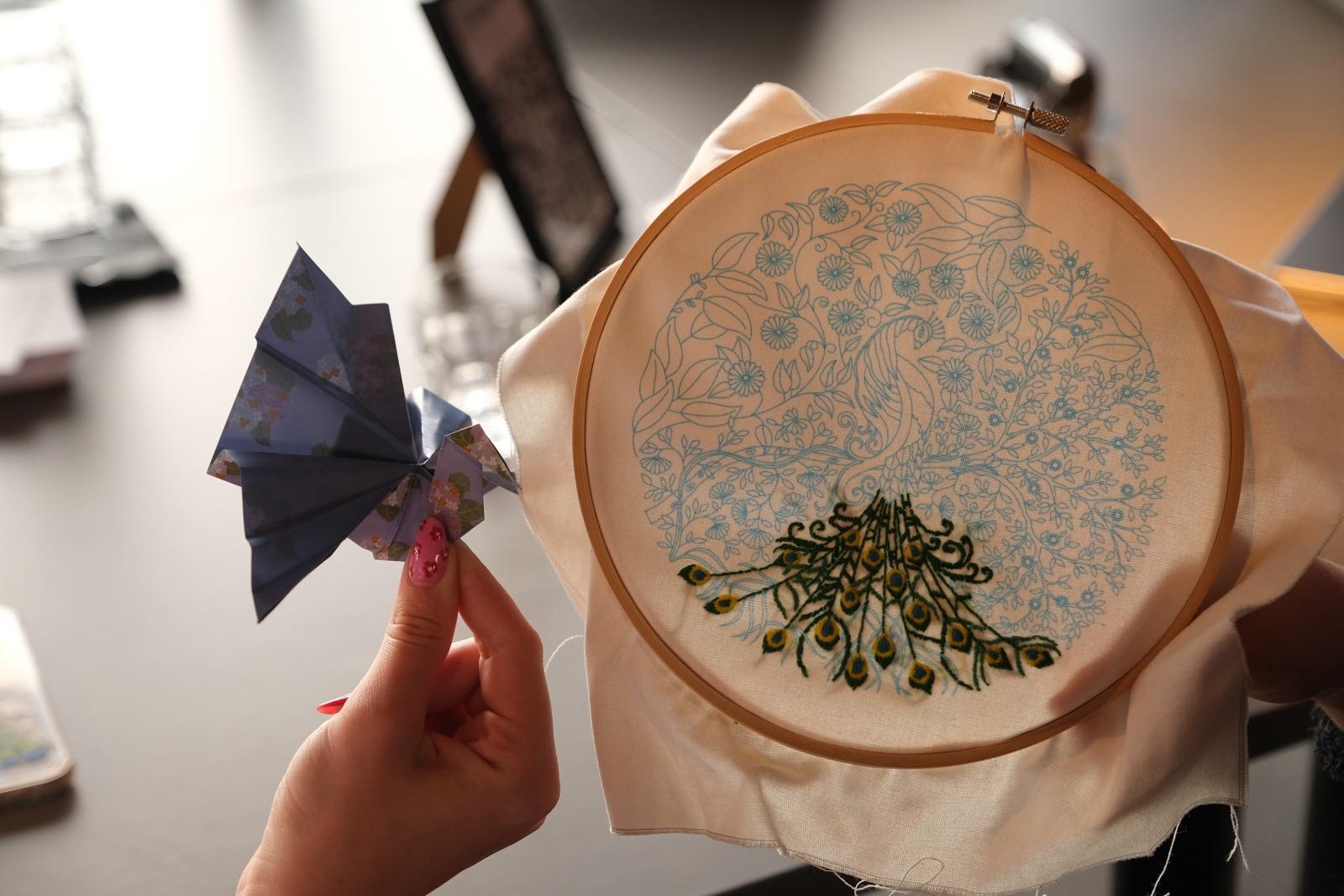 An origami crane held up next to an embroidery hoop with a stitched peacock